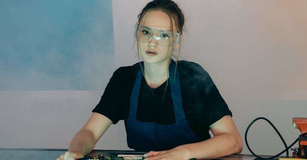 Woman in apron and goggles assembling electronic hardware indoors.