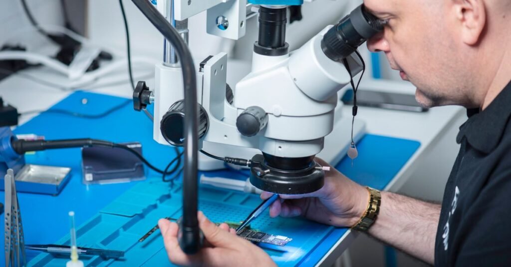 Technician examining electronic chip under a microscope in a computer lab setting.