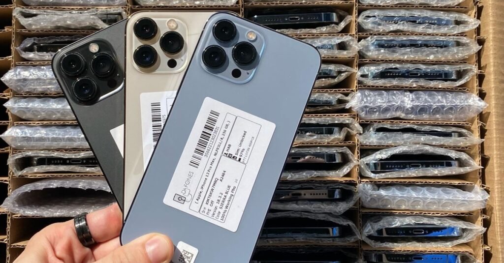 A hand holds three new smartphones over a box filled with packaged devices ready for shipment.